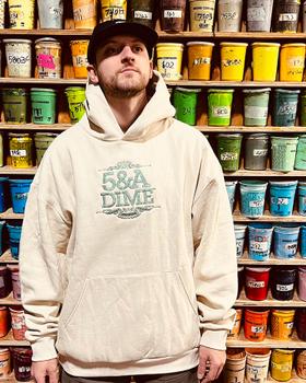 A man wearing a cream 5&A Dime embroidered hoodie and black cap in front of a wall of screen-printing ink buckets