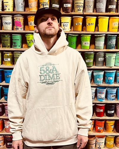 A man wearing a cream 5&A Dime embroidered hoodie and black cap in front of a wall of screen-printing ink buckets