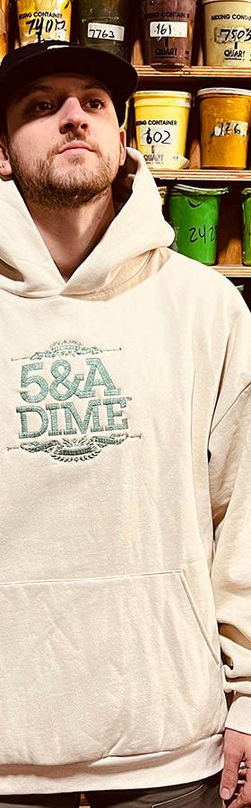 A man wearing a cream 5&A Dime embroidered hoodie and black cap in front of a wall of screen-printing ink buckets