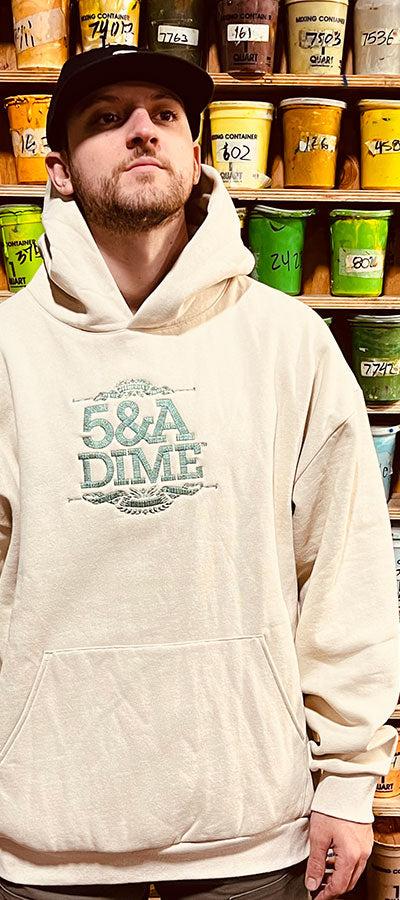 A man wearing a cream 5&A Dime embroidered hoodie and black cap in front of a wall of screen-printing ink buckets