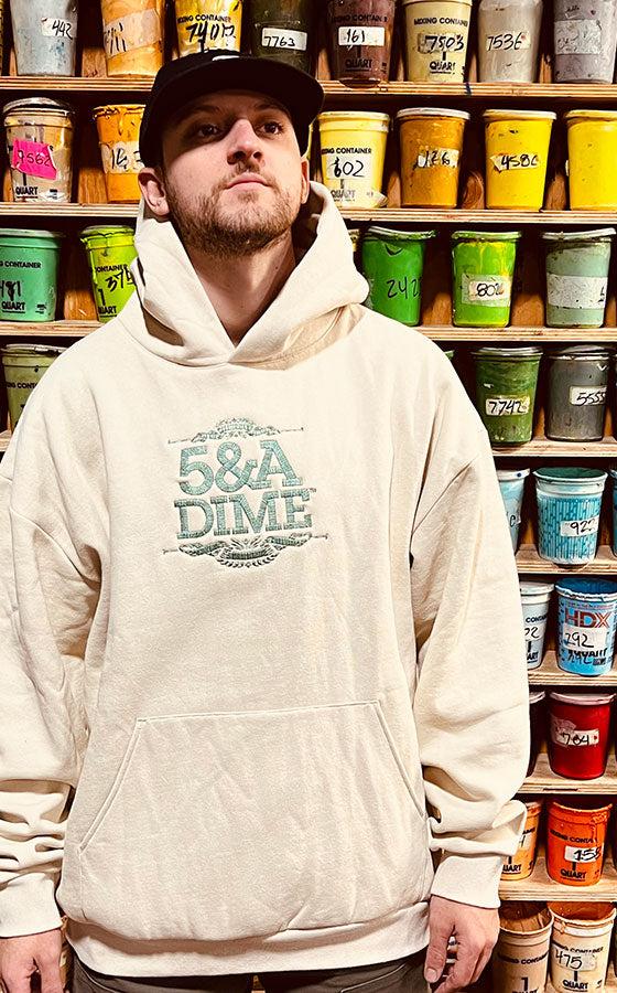 A man wearing a cream 5&A Dime embroidered hoodie and black cap in front of a wall of screen-printing ink buckets