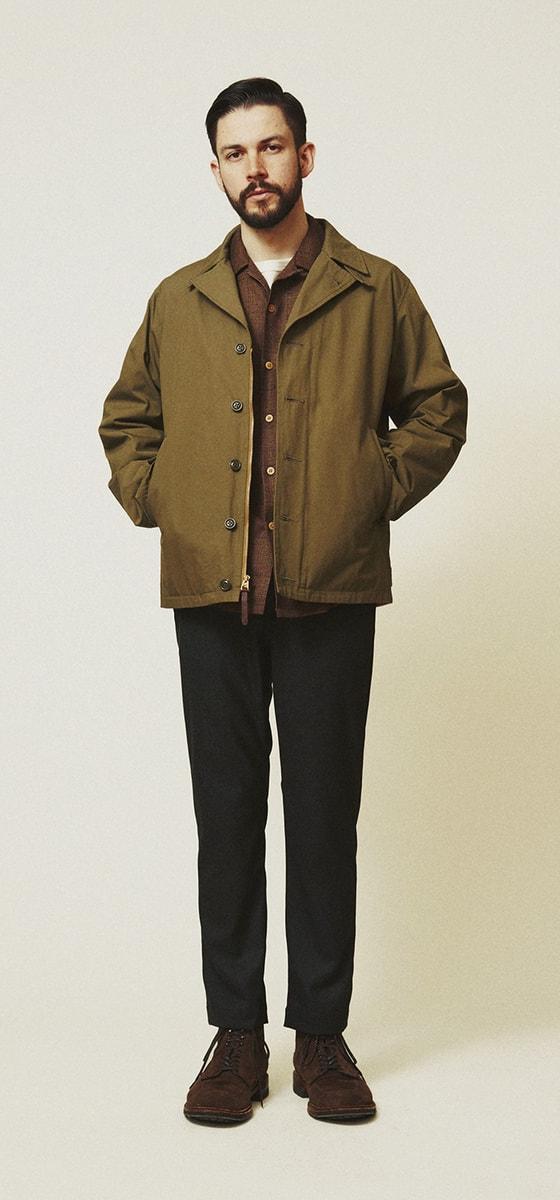 A man wearing an olive A VONTADE military deck jacket over a brown shirt with black trousers