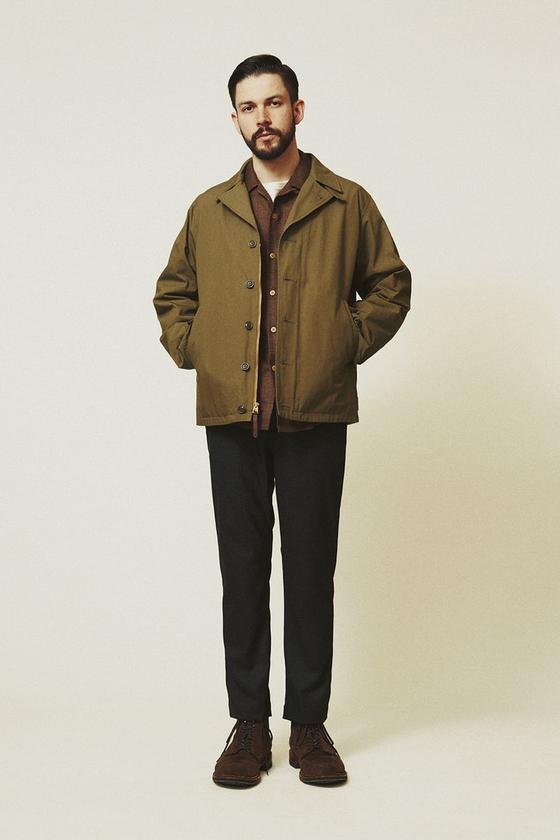 A man wearing an olive A VONTADE military deck jacket over a brown shirt with black trousers