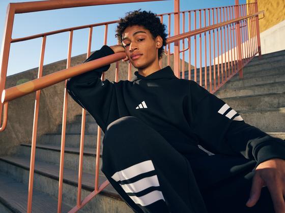 A man in a black adidas hoodie and three-stripe track pants sitting on concrete steps