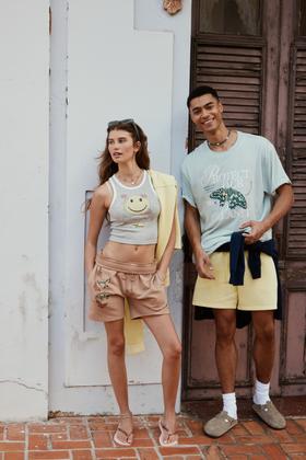 A woman in a cropped graphic tank and tan drawstring shorts standing beside a man in a pale blue printed tee and yellow shorts