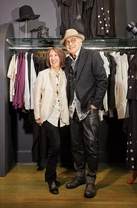 A couple wearing an ivory Alan Bilzerian jacket and a black blazer with leather trousers inside the boutique