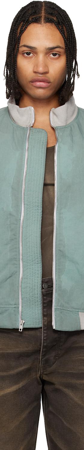 A man wearing a green Alexander DiGenova bomber jacket with faded brown jeans