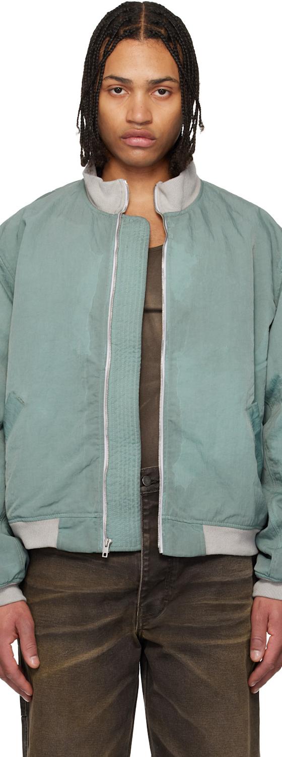 A man wearing a green Alexander DiGenova bomber jacket with faded brown jeans