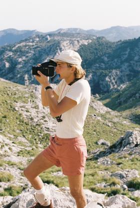 A man wearing a cream Amundsen Sports ribbed tee, coral shorts and a cap while filming in the mountains