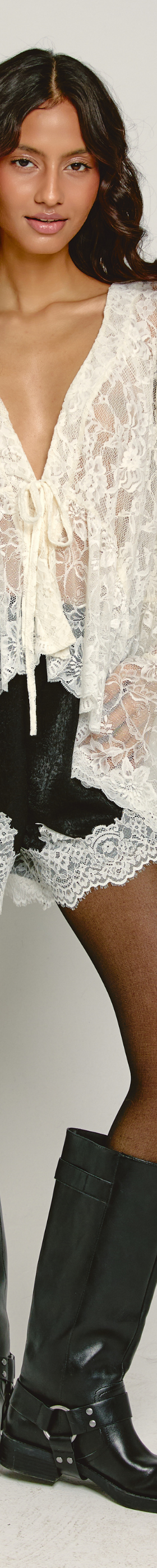 A woman wearing a sheer cream lace blouse and shorts with tall black leather boots