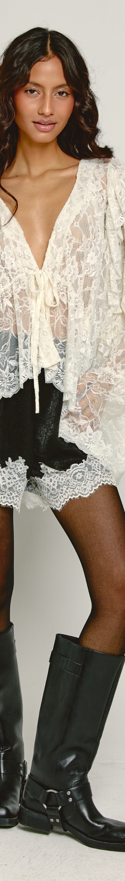 A woman wearing a sheer cream lace blouse and shorts with tall black leather boots