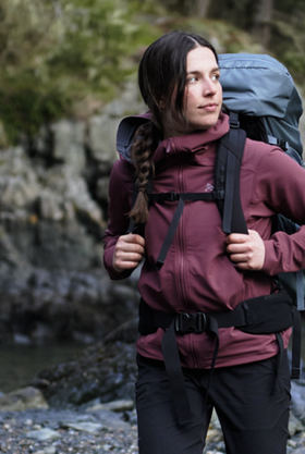 A woman wearing a burgundy Arc'teryx softshell jacket with a hiking backpack on a rocky shoreline
