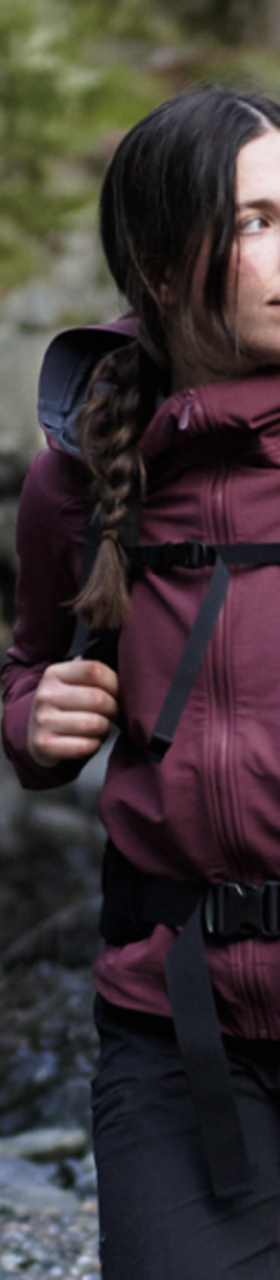 A woman wearing a burgundy Arc'teryx softshell jacket with a hiking backpack on a rocky shoreline
