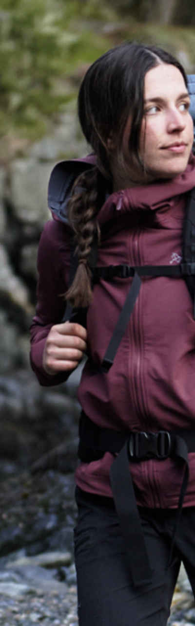 A woman wearing a burgundy Arc'teryx softshell jacket with a hiking backpack on a rocky shoreline