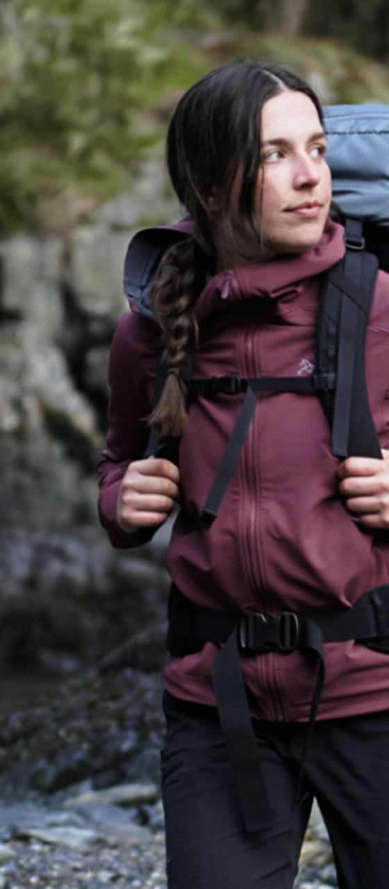 A woman wearing a burgundy Arc'teryx softshell jacket with a hiking backpack on a rocky shoreline