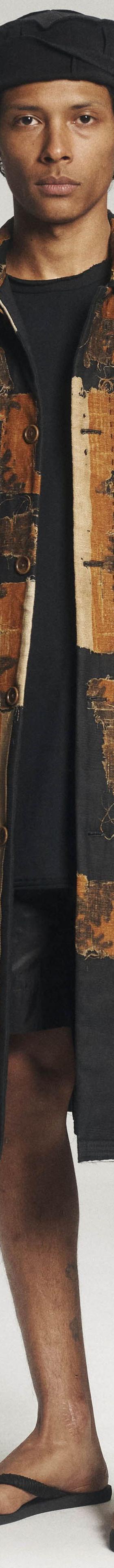 A man wearing a patchwork antique-textile Aubero coat with black shorts and sandals