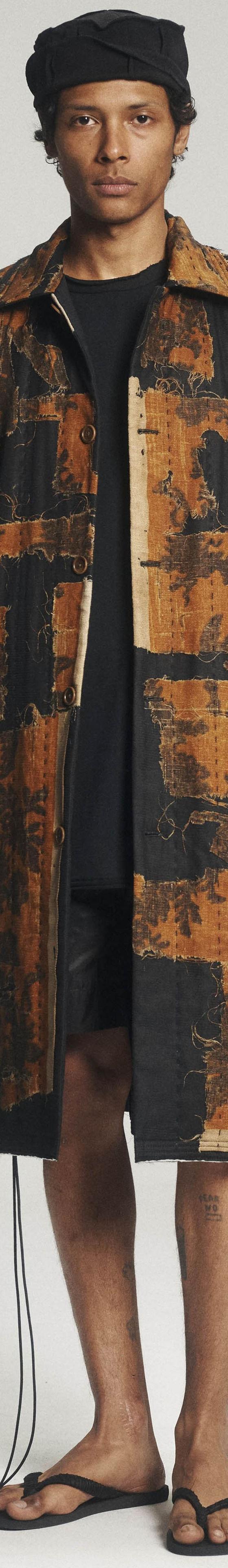 A man wearing a patchwork antique-textile Aubero coat with black shorts and sandals