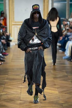 A man wearing an oversized black AWGE bomber jacket, distressed denim skirt-over-trousers and a printed cap on the runway