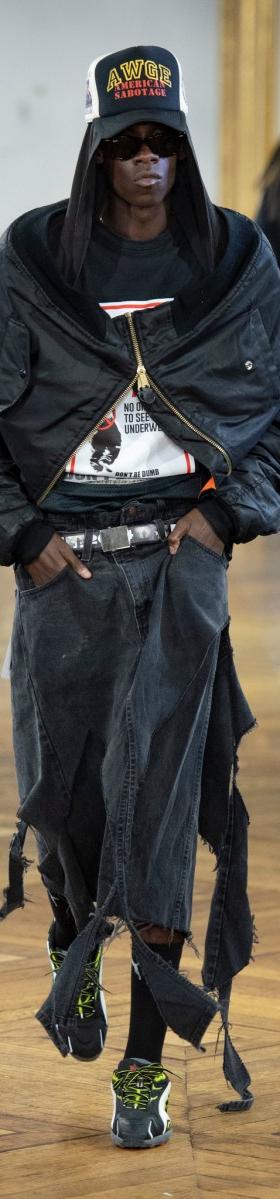 A man wearing an oversized black AWGE bomber jacket, distressed denim skirt-over-trousers and a printed cap on the runway