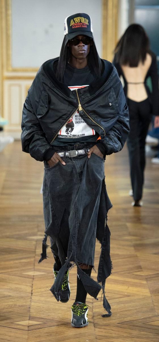 A man wearing an oversized black AWGE bomber jacket, distressed denim skirt-over-trousers and a printed cap on the runway