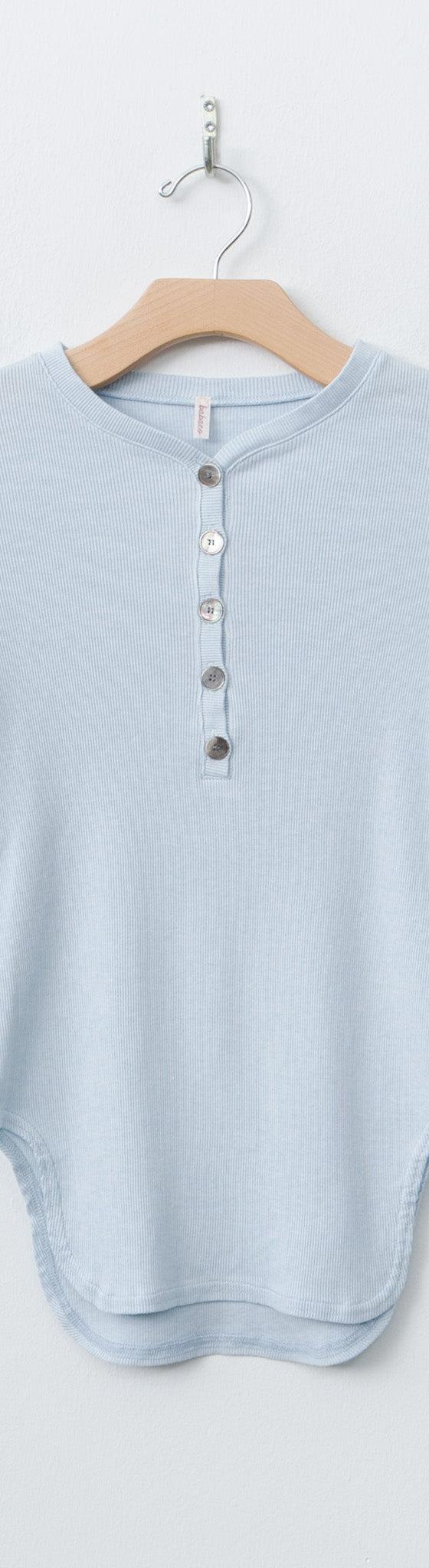 A pale blue babaco long-sleeve henley top on a wooden hanger against a white wall