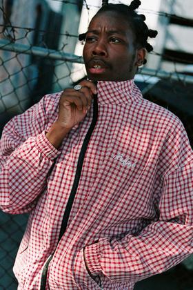 A man wearing a red and white checked Badfriend zip-up track jacket