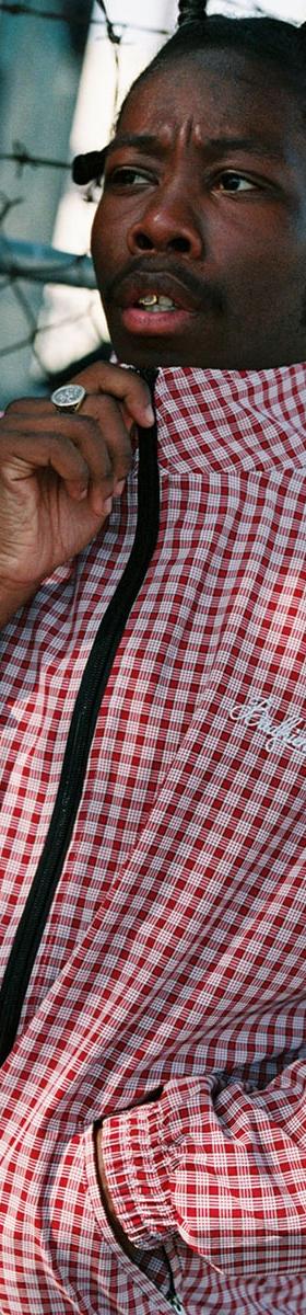 A man wearing a red and white checked Badfriend zip-up track jacket
