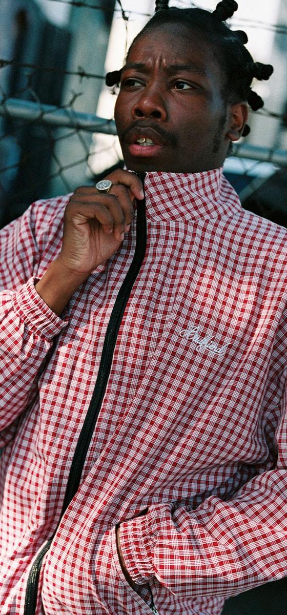 A man wearing a red and white checked Badfriend zip-up track jacket