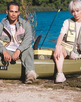 Two models in BAIT fishing-inspired jackets, vests and sneakers seated on a kayak by a lake