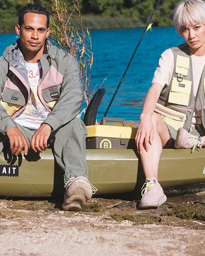 Two models in BAIT fishing-inspired jackets, vests and sneakers seated on a kayak by a lake