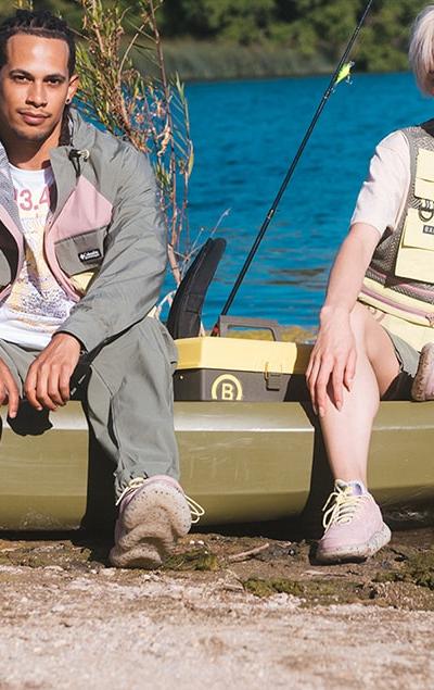 Two models in BAIT fishing-inspired jackets, vests and sneakers seated on a kayak by a lake