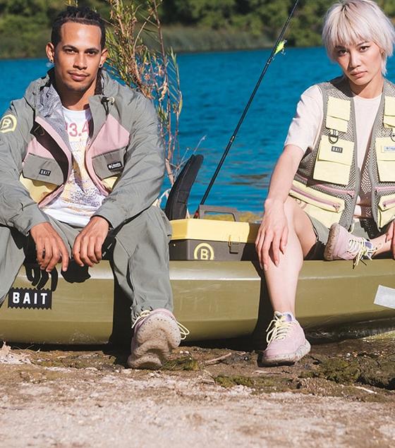 Two models in BAIT fishing-inspired jackets, vests and sneakers seated on a kayak by a lake