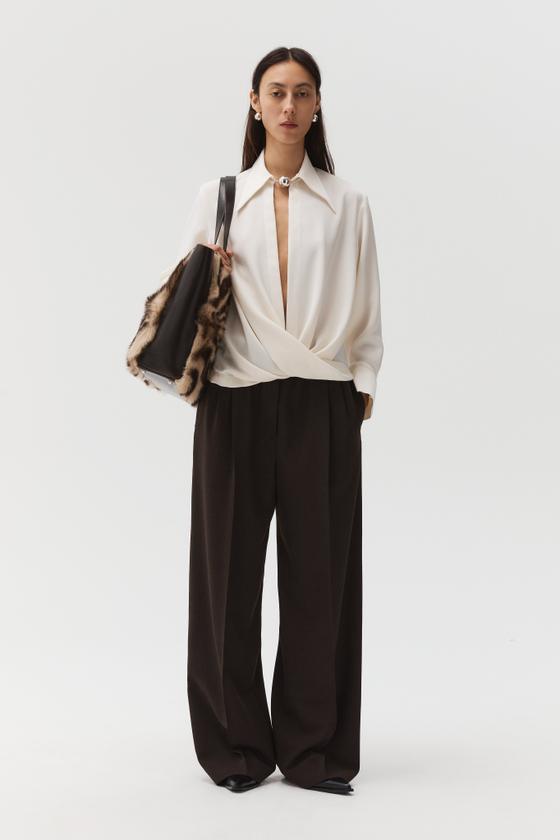 A woman wearing a white draped Beaufille blouse with dark brown wide-leg trousers
