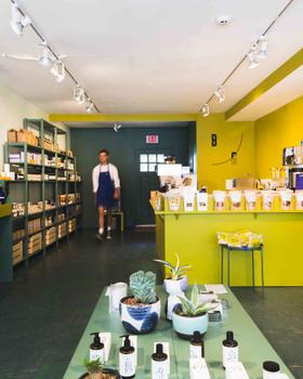 Interior of the Bellport General shop with shelves of provisions, a yellow coffee counter, and an aproned staff member