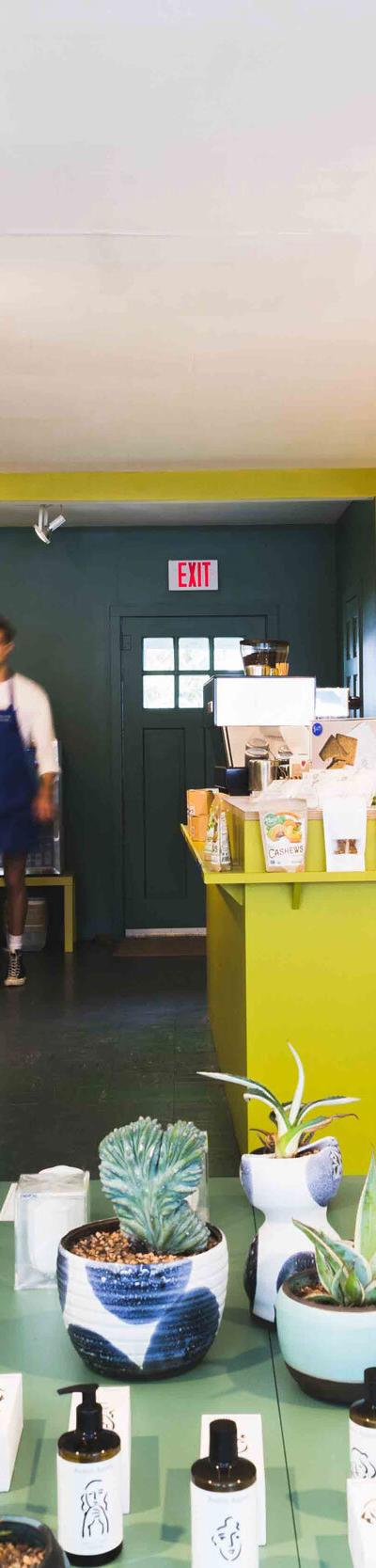 Interior of the Bellport General shop with shelves of provisions, a yellow coffee counter, and an aproned staff member