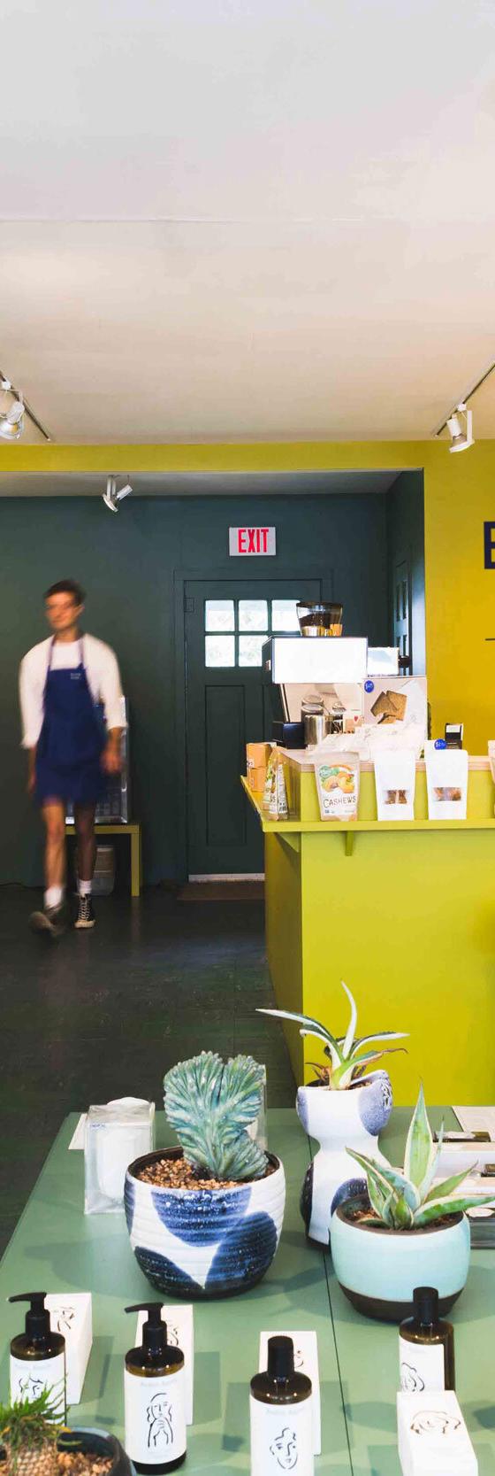 Interior of the Bellport General shop with shelves of provisions, a yellow coffee counter, and an aproned staff member