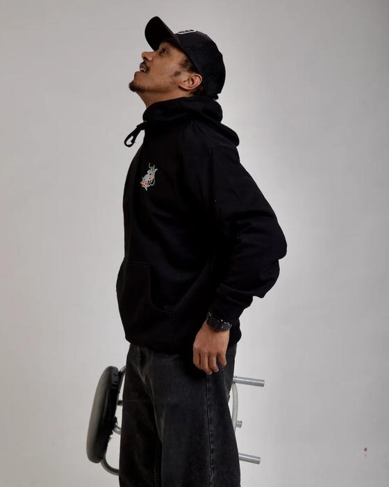 A man wearing a black Black Coffee Apparel hoodie with a small embroidered chest graphic, black jeans and a black trucker cap
