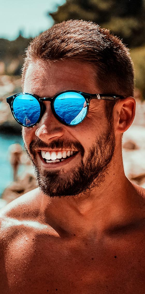 A man wearing blue mirrored Blenders Eyewear polarized sunglasses by the sea