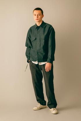 A man wearing a dark green blurhms overshirt with grey wide-leg trousers