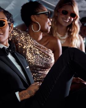 A group of people in tailored black and gold eveningwear with sunglasses, seated together in a car interior