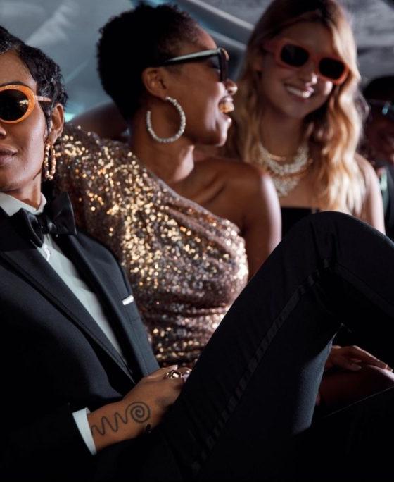 A group of people in tailored black and gold eveningwear with sunglasses, seated together in a car interior