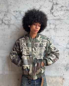 A person wearing a camo Bravest Studios bomber jacket with photo patches