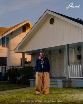 A man wearing a navy Bricks & Wood fleece pullover with a printed scarf and tan trousers in front of a South Central Los Angeles house
