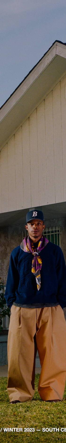 A man wearing a navy Bricks & Wood fleece pullover with a printed scarf and tan trousers in front of a South Central Los Angeles house