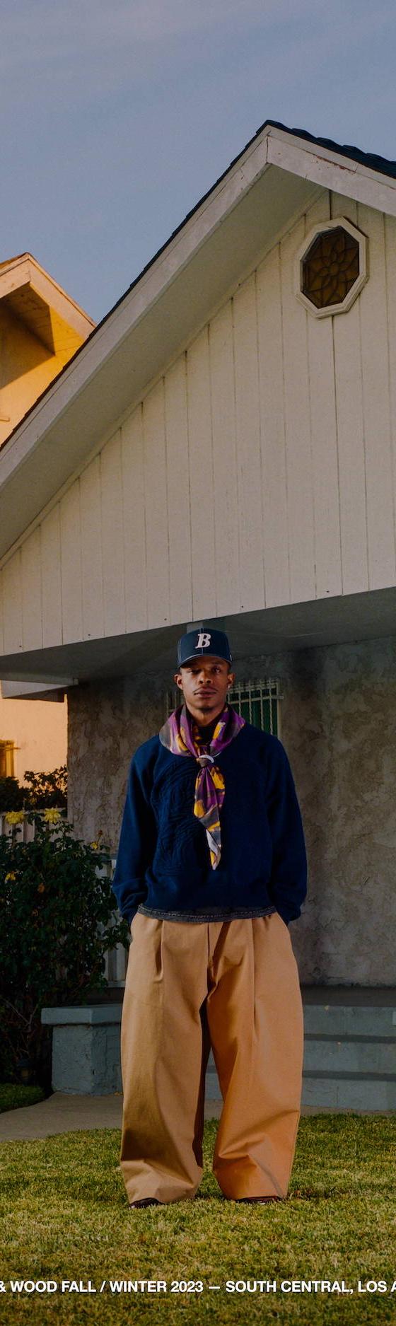 A man wearing a navy Bricks & Wood fleece pullover with a printed scarf and tan trousers in front of a South Central Los Angeles house
