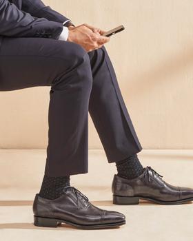 A man in a navy suit seated, wearing grey leather cap-toe Oxford shoes