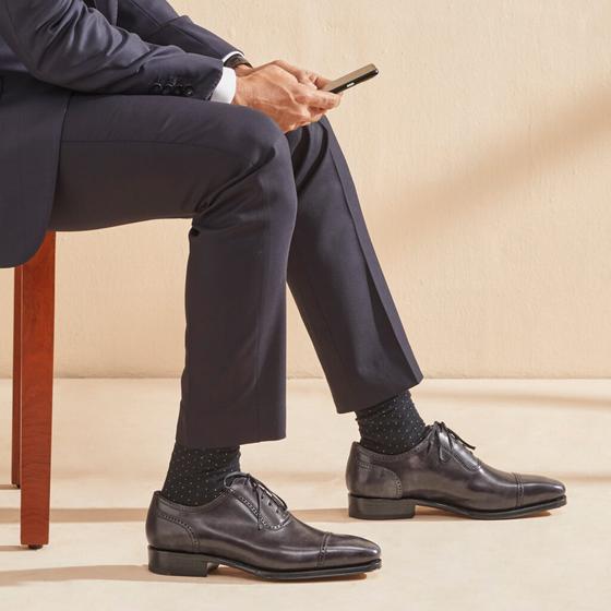 A man in a navy suit seated, wearing grey leather cap-toe Oxford shoes