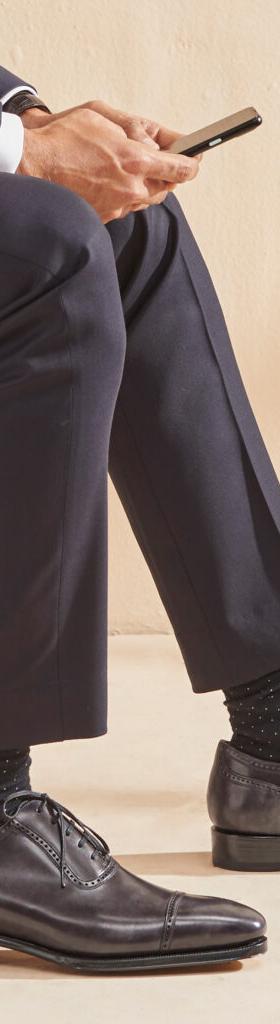 A man in a navy suit seated, wearing grey leather cap-toe Oxford shoes