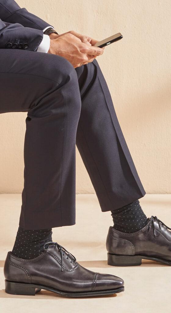 A man in a navy suit seated, wearing grey leather cap-toe Oxford shoes