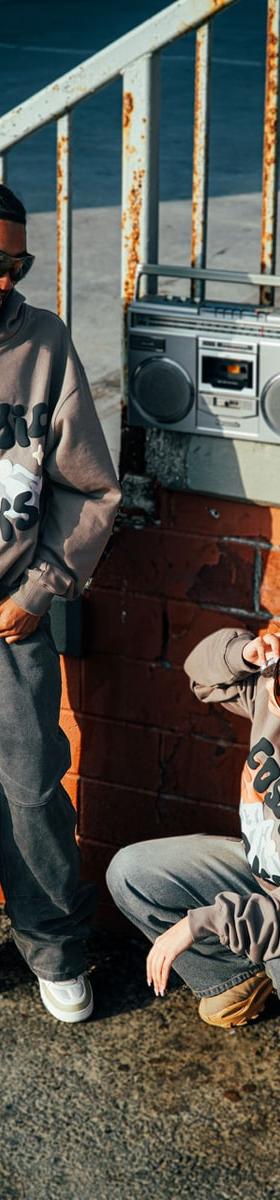 A man and a woman wearing taupe Broken Planet Market Cosmic Peaks hoodies and grey tracksuit bottoms beside an orange brick wall